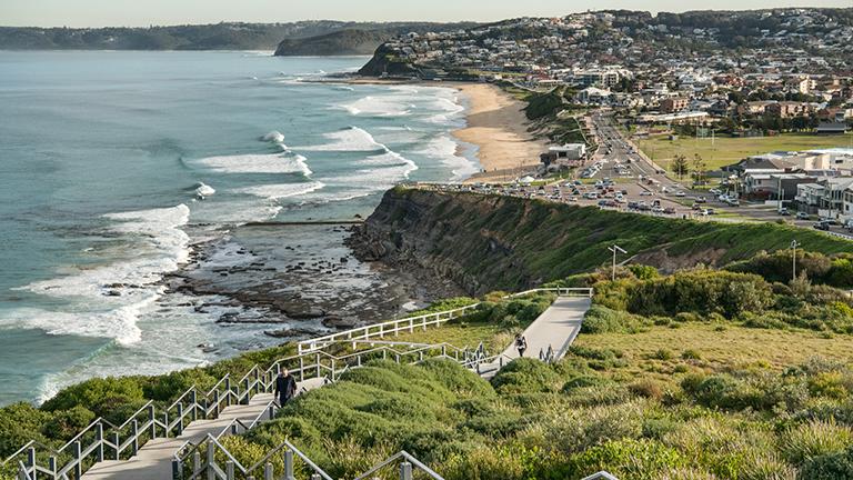 View of Newcastle coastline taken from high point on Newcastle Memorial Walk, featuring rolling ocean waves on left, lush grassy hill in foreground and cliffs and houses in distance.