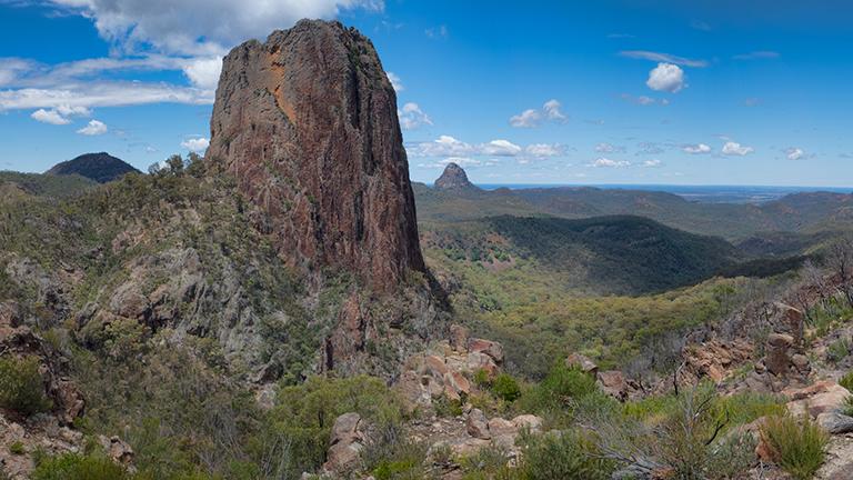 View south across Warrumbungle National Park from the Grand High Tops walking track, with large cylindrical Crater Bluff in the foreground and prominent rocky peak Tonduron Spire in the distance.