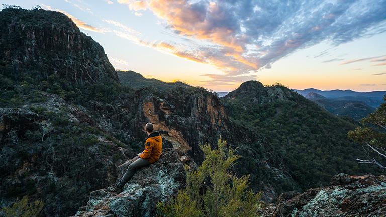 Man sitting on top of rock looking at beautiful Warrumbungles view at sunset.
